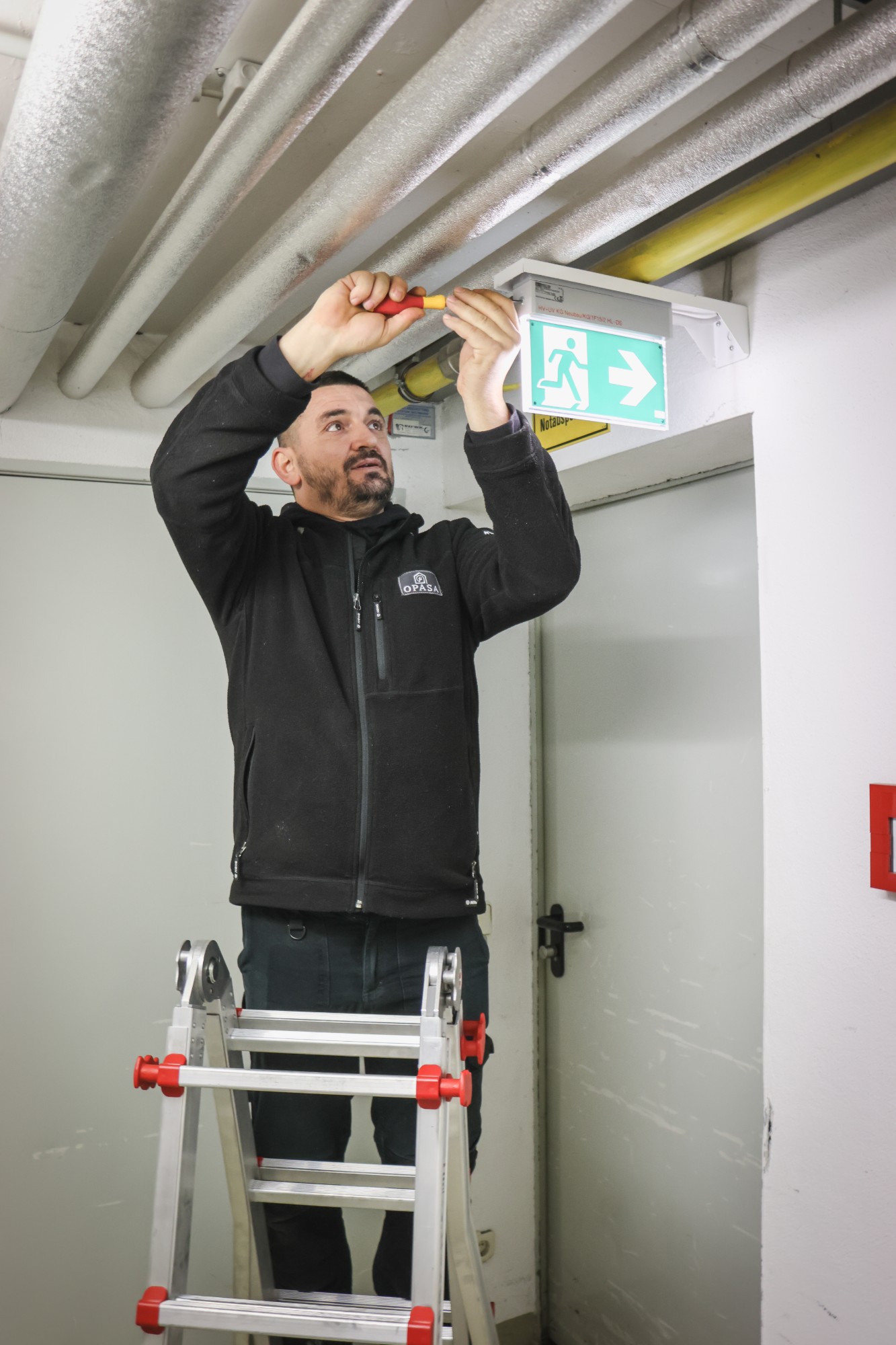 OPASA Elektrofachkraft montiert auf der Leiter eine Rettungszeichenleuchte